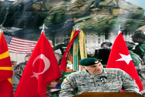 Ein Soldat steht an einem Podium mit einem Mikrofon, mit verschiedenen Flaggen und einem Plakat mit Militärpersonal im Hintergrund.