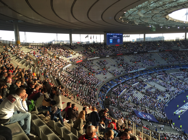 Große Zuschauermenge in einem Stadion bei einem Fußballspiel mit einer Bühne, Fahnen und einem Bildschirm im Hintergrund.