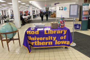 Eine Ausstellung mit einem Tisch, auf dem verschiedene Landesflaggen, eine Box, Papiere und eine Fahne sowie links eine Weltkugel, rechts ein Anschlagbrett mit einigen Menschen in der Nähe und viele Tische und Stühle zu sehen sind.