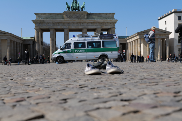 Ein Polizeiwagen steht vor dem Brandenburger Tor in Berlin, Deutschland, umgeben von Menschen und Fahrrädern, Schuhen auf dem Boden und Gebäuden, Säulen, Statuen, Bäumen und einem klaren blauen Himmel im Hintergrund.