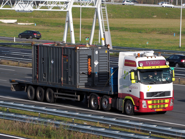 Ein Lkw mit einem großen Behälter fährt auf einer Autobahn, mit anderen Fahrzeugen, Masten, Bäumen, Gebäuden und einem klaren blauen Himmel im Hintergrund.