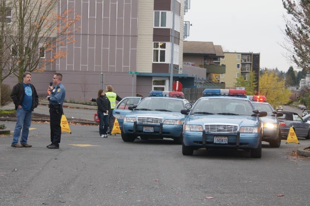 Autos auf einer Straße mit vier Personen in der Nähe, Gebäude mit Fenstern im Hintergrund, Bäume und was auszuscheiden scheinen Warnkegel.