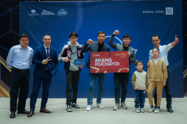 Eine Gruppe von Menschen auf einer Bühne mit einer Tafel, auf der der Text 'Akang Kuchaydi - Zakovat Quiz' steht, mit einem Banner, Scheinwerfern und anderen Gegenständen im Hintergrund.