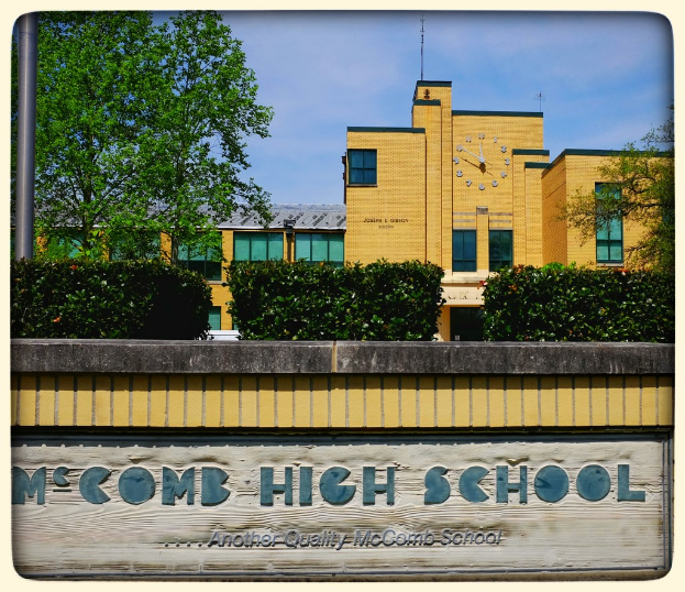 Ein Plakat mit dem Text "MCCOMB HIGH SCHOOL" auf einer Wand mit Pflanzen, Bäumen, einem Pfahl, einem Gebäude und einem bewölkten Himmel im Hintergrund.