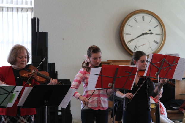 Drei Frauen, die auf der Bühne Geigen spielen, mit Notenpulten davor, einem im Vordergrund sitzenden Publikum, einer Uhr an der weißen Wand im Hintergrund und einem weißen Vorhang in der linken Ecke.