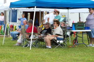 Eine Gruppe von Menschen, einige sitzen und einige stehen, versammelt sich an einem Outdoor-Standort mit Zelten, einem Tisch, Stühlen und einem Fahrzeug im Hintergrund. Einige Individuals tragen Goggles und es gibt dekorative Gegenstände auf der linken Seite, mit Körben und einem Konus auf dem Tisch.