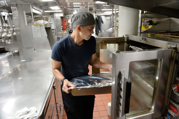 Ein Mann in Kochuniform steht in einer Küche und hält ein Tablett mit Essen vor einem offenen Ofen, mit Utensilien an der linken Seite und einem Herd mit verschiedenen Gegenständen auf der rechten Seite.