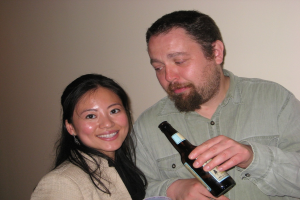 Ein Mann hält eine Flasche, während er eine lächelnde Frau ansieht, beide stehen vor einer Wand.