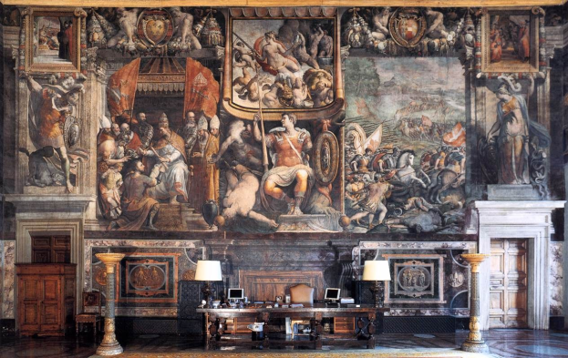 Ein großer Raum im Palazzo della Signoria in Florenz, Italien, mit einem großen Gemälde an der Wand, einem Tisch mit verschiedenen Gegenständen, zwei Lampen, zwei Türen und einem Teppich.