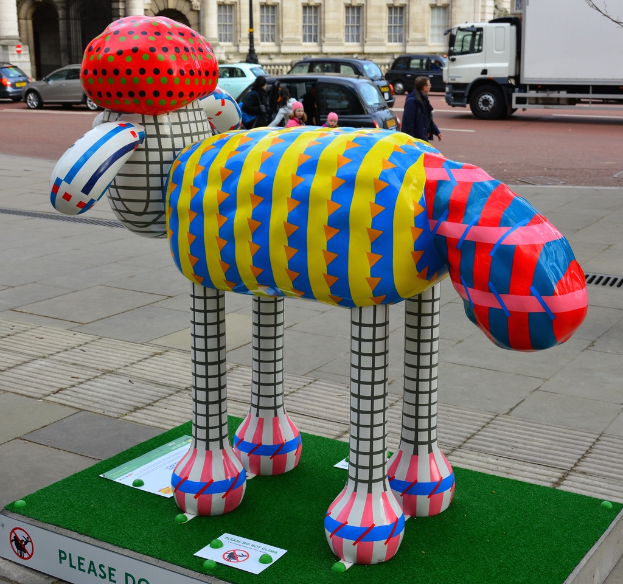 Eine Skulptur auf dem Boden mit Brettern in der Nähe, Fahrzeuge auf der Straße dahinter, einige Menschen auf einem Gehweg und Gebäude mit einem Mast im Hintergrund.