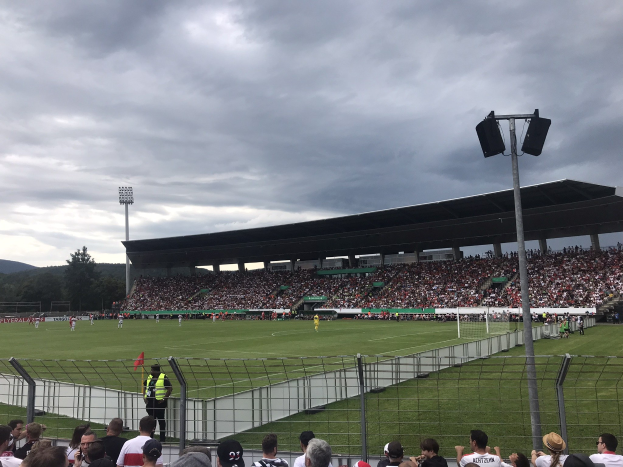 Große Zuschauermenge bei einem Fußballspiel in einem Stadion mit beleuchtetem Feld, eingezäuntem Umfang und Bäumen und Hügeln im Hintergrund bei bewölktem Himmel.