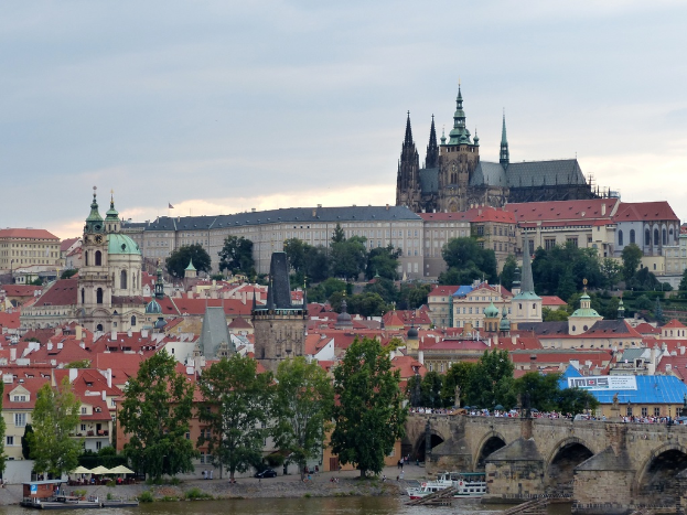 Ein Schiff auf dem Wasser mit einer Brücke und Menschen dahinter, mit Bäumen, Gebäuden, einem Schloss und dem Himmel im Hintergrund.