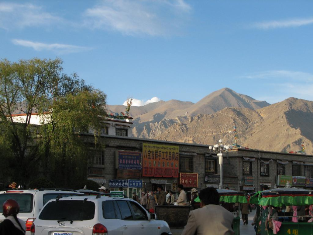Fahrzeuge, Menschen, Zelte und Stangen auf einer Straße mit Bäumen, Gebäuden und Bergen im Hintergrund.
