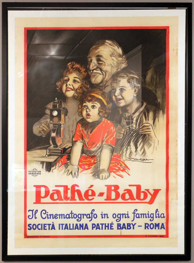 Ein Foto Rahmen an der Wand zeigt ein Poster einer Familie mit drei Personen und dem Text "Pathé-Baby - Il Cinematografico in Ogni Famiglia Società Italiana Pathé Baby - Roma."