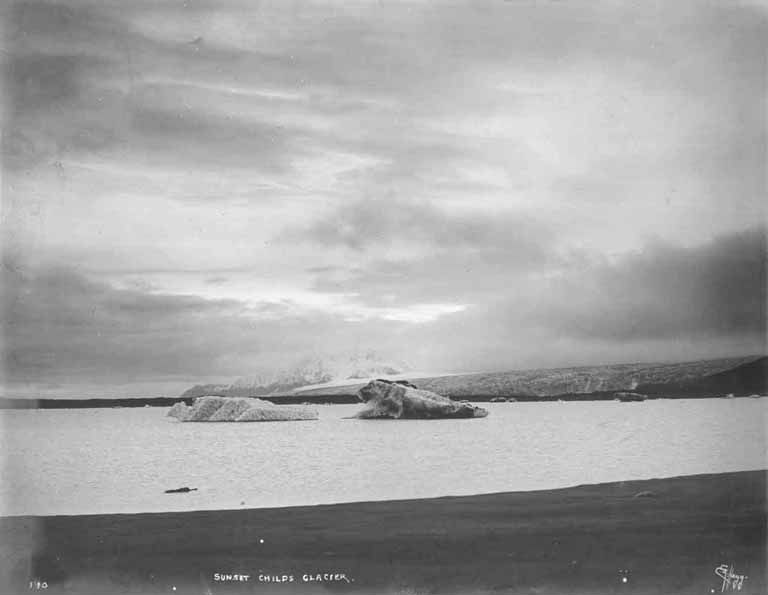 Ein Schwarz-Wei├č-Foto eines Eisbergs, der im Ozean treibt, umgeben von H├Ąngen und einem bew├Âlktem Himmel, mit dem Text "Sunset Childs Glacier" unten.