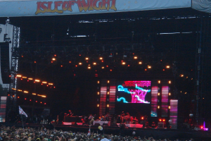 Eine Bühne mit Lichtern, einem Bildschirm und Musikern, die Instrumente spielen, mit einem Banner oben, einer Menge unten und Fahnen mit Stangen und Ballons.