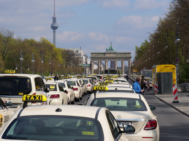 Eine belebte Berliner Straße mit zahlreichen parkenden Taxis, Fußgängern auf dem Gehweg, gesäumt von Laternenmasten, Bäumen und Gebäuden, mit einem fernen Bogen mit Statuen und einem Turm unter einem bewölkten Himmel.