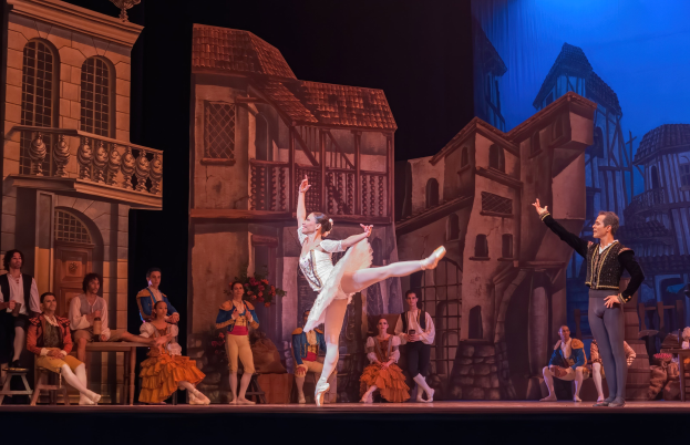 Zwei Tänzer performen auf einer Bühne im Royal Opera House in London, mit einem Publikum im Vordergrund und einer Anzeige mit Gebäudeabbildungen im Hintergrund.