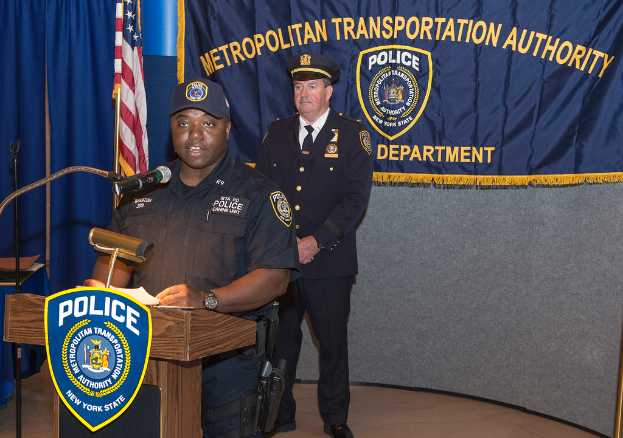 Zwei Polizisten stehen vor einem Rednerpult, einer hölt Papiere und spricht in ein Mikrofon, mit einer Fahne, einem Tisch und einer Tafel mit Symbolen und Text im Hintergrund.