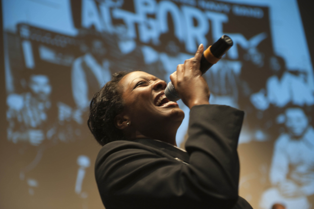 Eine Frau in einem schwarzen Kleid singt leidenschaftlich in ein Mikrofon auf der Bühne, ihre Augen geschlossen und ein intensiver Ausdruck im Gesicht, mit Scheinwerfern, die ihr Gesicht beleuchten.