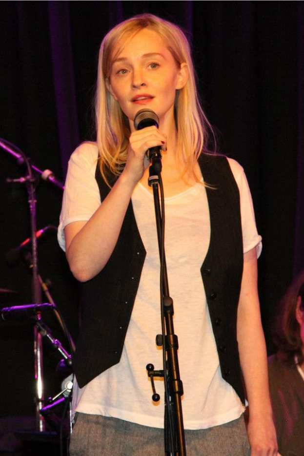 Eine Frau in einem weißen T-Shirt und schwarzer Jacke hält ein Mikrofon in der Hand und steht auf einer Bühne mit einer weiteren Person rechts von ihr, umgeben von Mikrofonen und Musikinstrumenten.