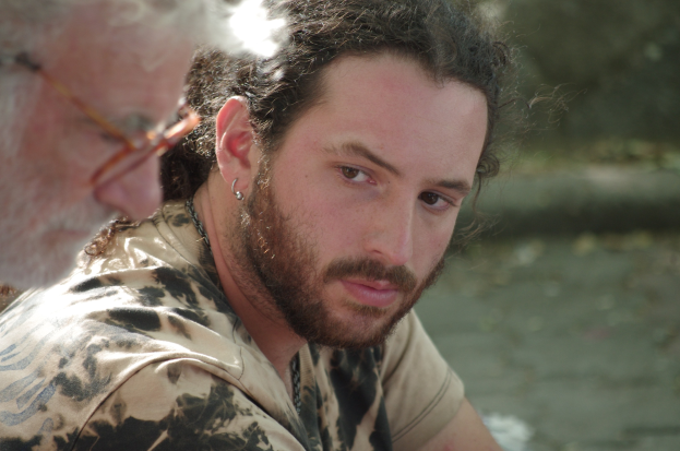 Zwei Männer sitzen in der Nähe eines Gewässers, einer trägt eine Brille, mit leicht unscharfem Hintergrund.