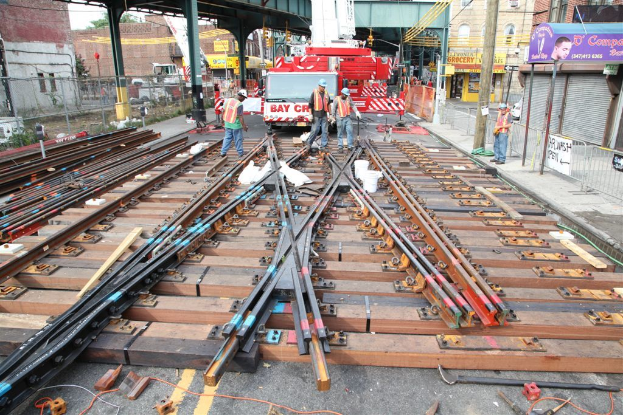 Arbeiter in Jacken und Mützen bauen Schienen, mit einem Fahrzeug, das mit Materialien beladen ist, und einer Hütte mit Pfählen im Hintergrund und einem Geschäft auf der anderen Seite.