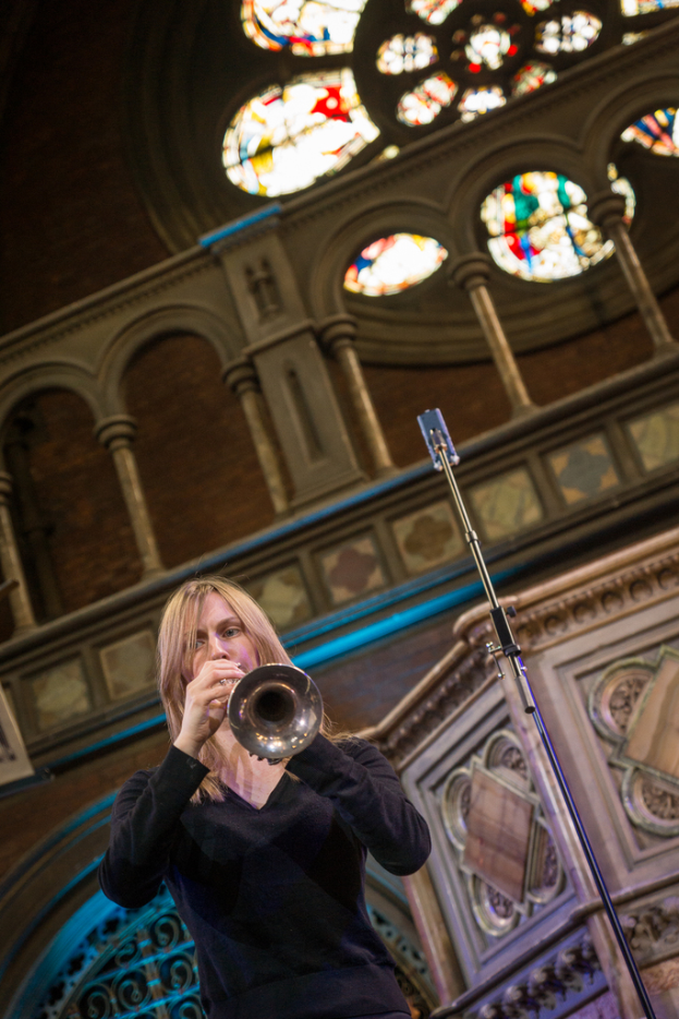 Eine Frau in einem schwarzen Kleid spielt eine Trompete neben einem Mikrofon, mit einem Gebäude und einem Geländer dahinter und dem Dach des Gebäudes oben.