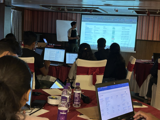 Eine Gruppe von Menschen, die um einen Tisch mit Laptops, Flaschen, Papieren und Stiften sitzen und an einem Businessplan-Training teilnehmen, während im Hintergrund ein Referent mit einem Mikrofon steht.