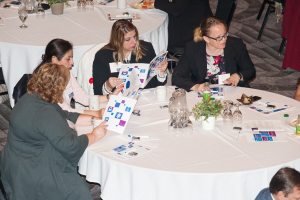 Vier Frauen auf Stühlen sitzend, eine hält ein Buch, mit einem Tisch davor, der einen Krug, Gläser, eine Tasse, einen Blumentopf, eine Pflanze, einen Teller und eine Flasche sowie mehrere andere Personen im Hintergrund zeigt.
