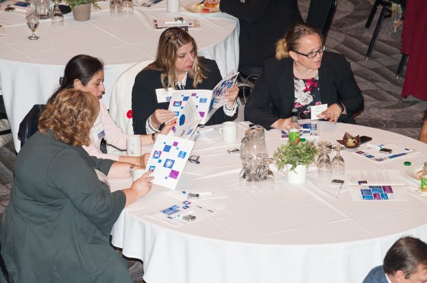 Vier Frauen auf Stühlen sitzend, eine hält ein Buch, mit einem Tisch davor, der einen Krug, Gläser, eine Tasse, einen Blumentopf, eine Pflanze, einen Teller und eine Flasche sowie mehrere andere Personen im Hintergrund zeigt.