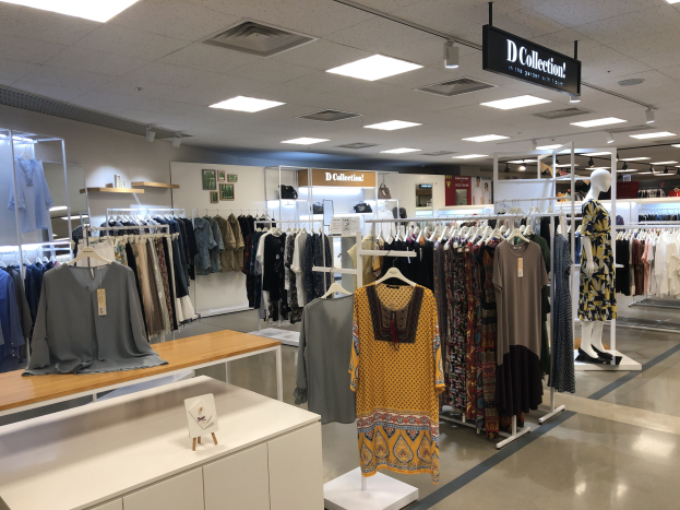 Innenansicht eines Kleiderladens mit Kleidern an Kleiderbügeln, Schaufensterpuppen, Tischen mit Auslagen, Wandtafeln und Deckenbeleuchtung, beschriftet als der neue D Collection Store im Mall of America.