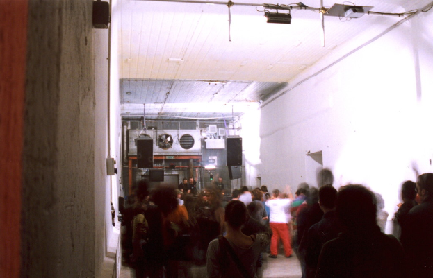 Leute tanzen in einem Raum mit Lautsprechern, einer Rohrleitung und einer weißen Wand, unter Lampen, einem Projektor und einem Eisenrahmen, mit verschiedenen Gegenständen im Hintergrund.