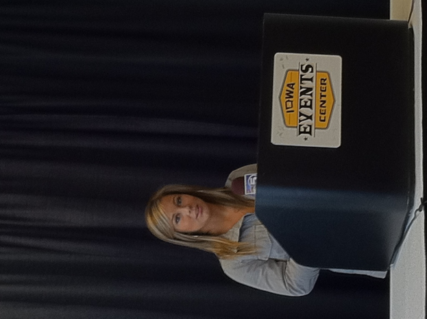 Eine Frau in einem blauen Hemd steht neben einem Podium mit Text darauf, mit einem Vorhang im Hintergrund.