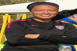 Zwei Frauen stehen auf einem Feld, eine hält eine Wasserflasche, mit einem Zelt, gelben Pfählen, Bäumen und einem klaren blauen Himmel im Hintergrund.