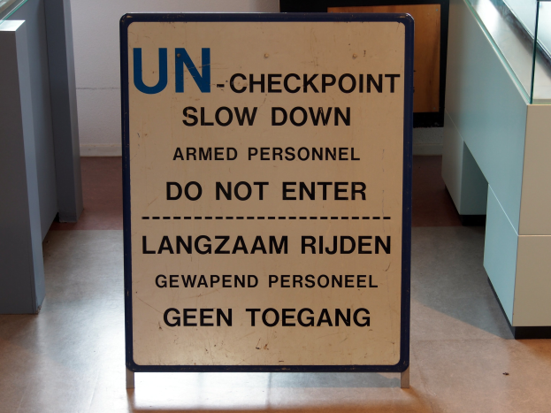 Schild auf dem Boden mit der Aufschrift "UN-Checkpoint Verlangsame bewaffnete Person - Betreten verboten" mit einem Glastisch und Gegenständen rechts und einer Wand im Hintergrund.