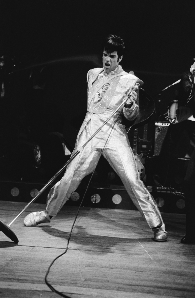 Elvis Presley in weißem Jumpsuit auf der Bühne des Apollo Theaters in Las Vegas, ein Mikrofon haltend, mit einem anderen Musiker rechts von ihm und Bühnenlichtern und Instrumenten im Hintergrund.