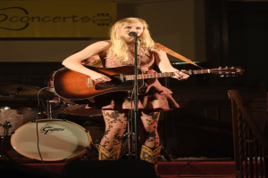 Eine Frau, die auf der Bühne Gitarre spielt und singt, mit einer Band im Hintergrund und einem Bildschirm an der Wand