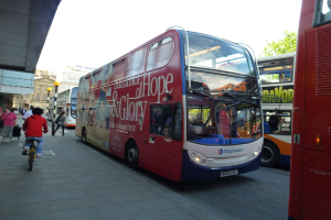 Eine Person f√§hrt ein Fahrrad auf einem Gehweg mit vielen Menschen, Bussen mit Text darauf, Geb√§uden und Laternen im Hintergrund.