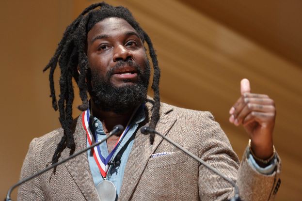 David Oyelowo, ein Mann mit Dreadlocks in einem Blazer und einer Medaille, hält eine Rede vor einem Rednerpult mit einem unscharfen Hintergrund.