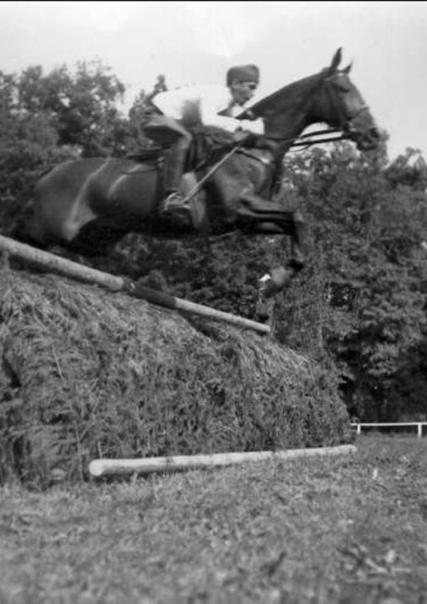 Ein Schwarz-Weiß-Foto eines kastanienbraunen Pferdes mit einer weißen Blesse auf der Stirn, das mittig über einen Strohballen springt, mit wehender Mähne und Schweif, während ein Reiter darauf sitzt, mit Bäumen und einem klaren blauen Himmel im Hintergrund.