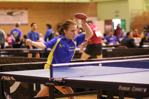 Eine Frau wird beim Tischtennis mit einem Schläger gezeigt, umgeben von ein paar sitzenden und stehenden Personen und einem Banner an der Wand.