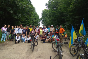 Eine Gruppe von Menschen steht neben ihren Fahrrädern auf einem Schotterweg, der von Bäumen umgeben ist und einen klaren blauen Himmel hat, einige tragen Helme und Taschen.