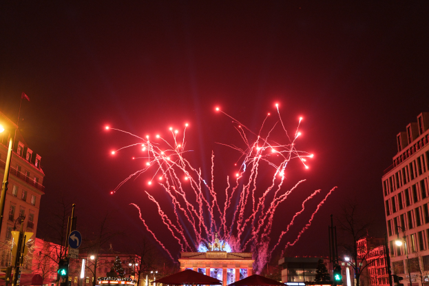 Eine Straße in Berlin an Silvester, voller Gebäude, Bäume, Laternenmäste, Verkehrszeichen, Zelte und Menschen, mit einem von Feuerwerk erhellten Himmel im Hintergrund.