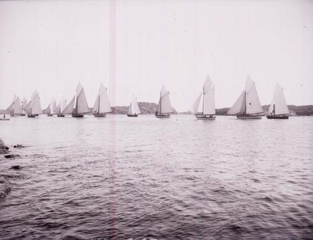 Schwarzes und weißes Foto von Segelbooten auf dem Wasser mit Hügeln im Hintergrund und einem klaren Himmel, als Foto des New York Yacht Clubs bezeichnet.