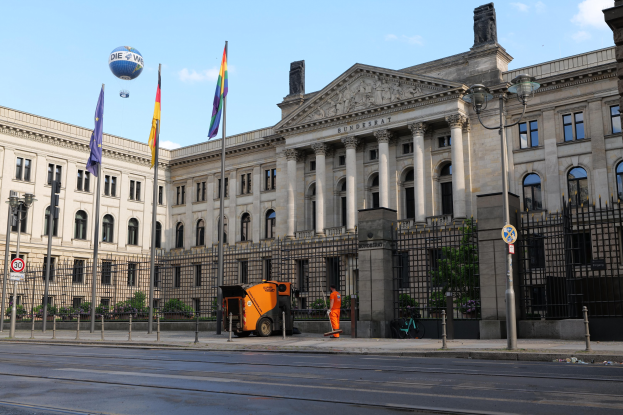 Großes modernes Gebäude mit zahlreichen Fenstern, als Bundestag in Berlin identifiziert, umgeben von Straßeninfrastruktur, Fußgängern und Bäumen unter einem bewölkten Himmel.