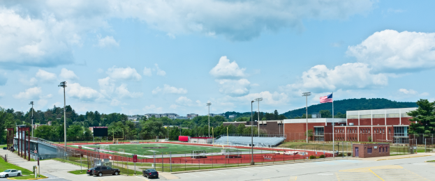 Ein Fußballfeld mit parkenden Autos im Vordergrund, umgeben von Gebäuden, Straßenmästen, Straßenlaternen, einer Flagge, Bäumen, Hügeln und einem bewölkten Himmel im Hintergrund.