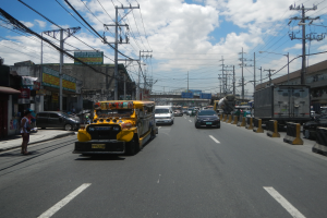 Ein gelber Schulbus fährt eine Straße mit hohen Gebäuden, Strommästen und Kabeln entlang, mit einigen Passanten auf dem Gehweg und einer bewölkten Himmel im Hintergrund.