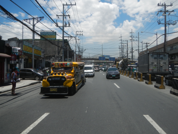 Ein gelber Schulbus fährt eine Straße mit hohen Gebäuden, Strommästen und Kabeln entlang, mit einigen Passanten auf dem Gehweg und einer bewölkten Himmel im Hintergrund.