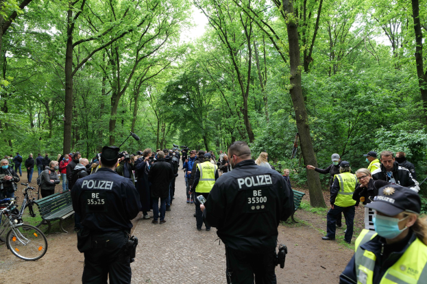 Gruppe von Polizisten vor einer Menschenmenge bei einer Outdoor-Veranstaltung, mit Fahrrädern und einer Bank im Vordergrund und Bäumen im Hintergrund.